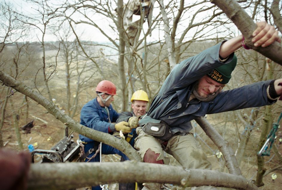 Newbury Bypass 1996 – 60 feet above the ground a man desperately attempts to hang on to the tree in order to protect it from being cut down whilst physically being pulled backwards by the Sheriff of Newbury’s eviction men.