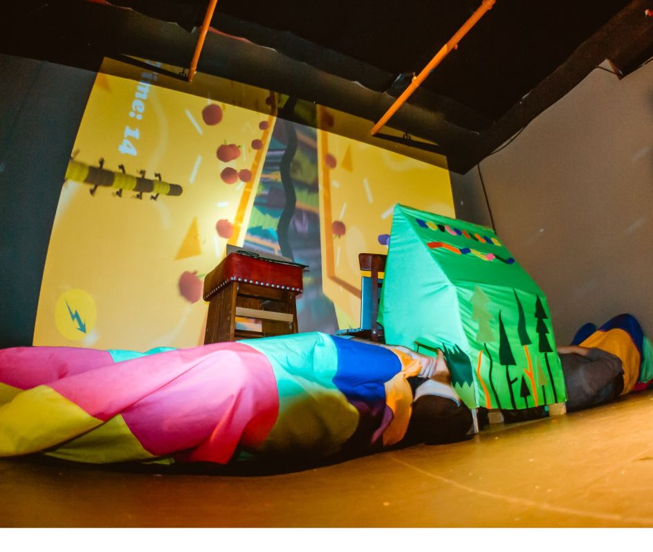 Roflpillar: Two players dress as rainbow-coloured caterpillars and wriggle around on the floor, competing to collect the most apples. The game's screen is fixed to the roof inside the fabric playhouse where players stick their heads. Image: Lucky Frame