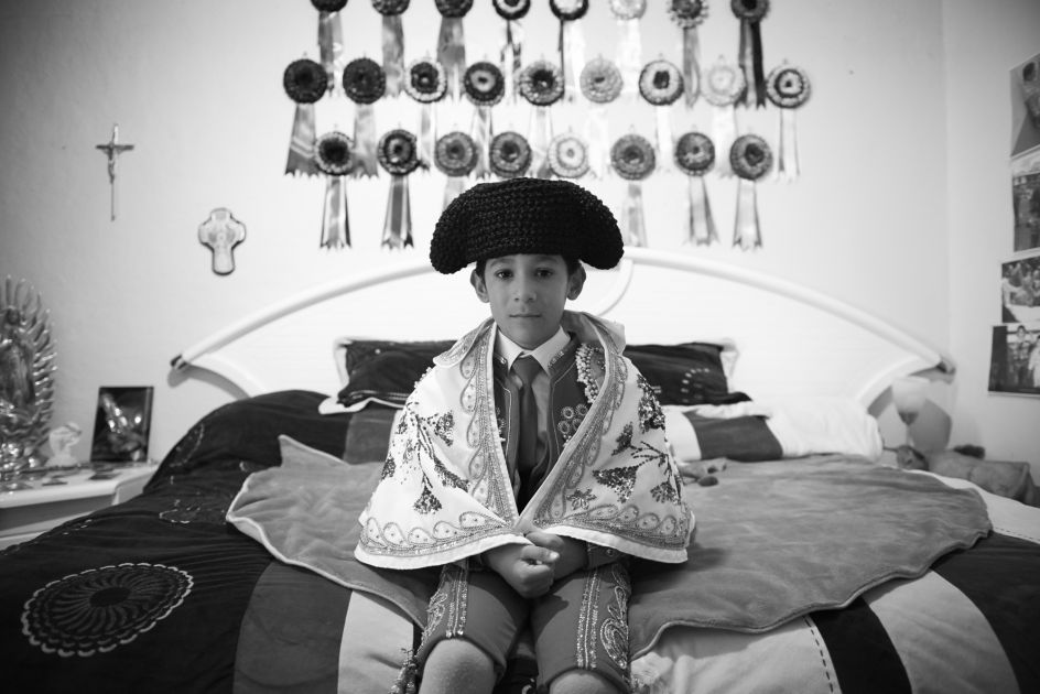 Salvador, aged 6, sits on his bed in anticipation of witnessing the afternoon bullfight at Plaza de Toros Mexico. Behind him are several ribbons, each symbolize a bull dedicated to him by his matador heroes. Salvador's family is wealthy and staunch Catholics. They believe God will protect him from any harm | © Christina Simons, Iceland, Shortlist, Professional, Daily Life, 2017 Sony World Photography Awards