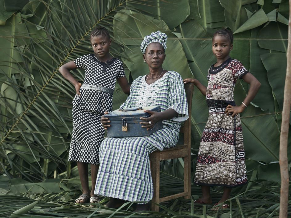 Mayama Mustafa, 60, women's leader and village midwife, with the two children she looks after: Jeneba, left, and Nafisatu, her niece. She holds the savings bank that all women contribute to and share out. Tombohuaun, Kailahun Distrcit, Sierra Leone, May 2017. 	WaterAid/ Joey Lawrence