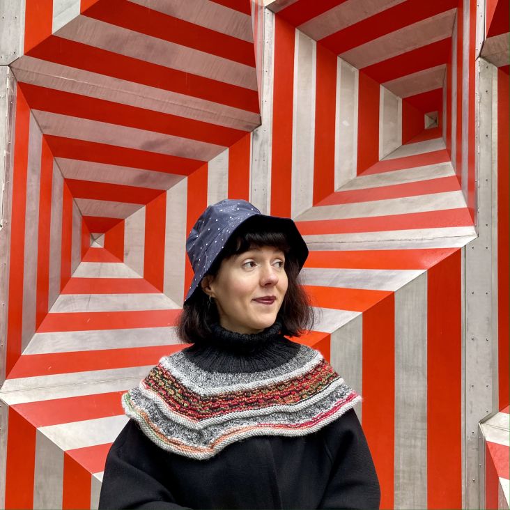 Hatiye (she/her/disabled), a medium light-skinned person with dark and short bob haircut, is in front of a red and gray background with geometric shapes reminiscent of op art. She wears a navy blue polka dot rain hat, looks to the left and smiles. She also wears a black poncho and hand-knit Icelandic collar.
