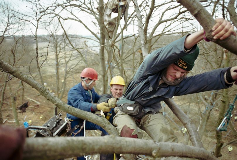 Newbury Bypass 1996 – 60 feet above the ground a man desperately attempts to hang on to the tree in order to protect it from being cut down whilst physically being pulled backwards by the Sheriff of Newbury’s eviction men.