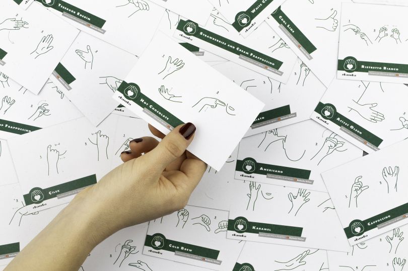 A photo of scattered cards in Turkish sign language, with a hand holding one of them. The cards show in a few steps how to sign coffee-related words.