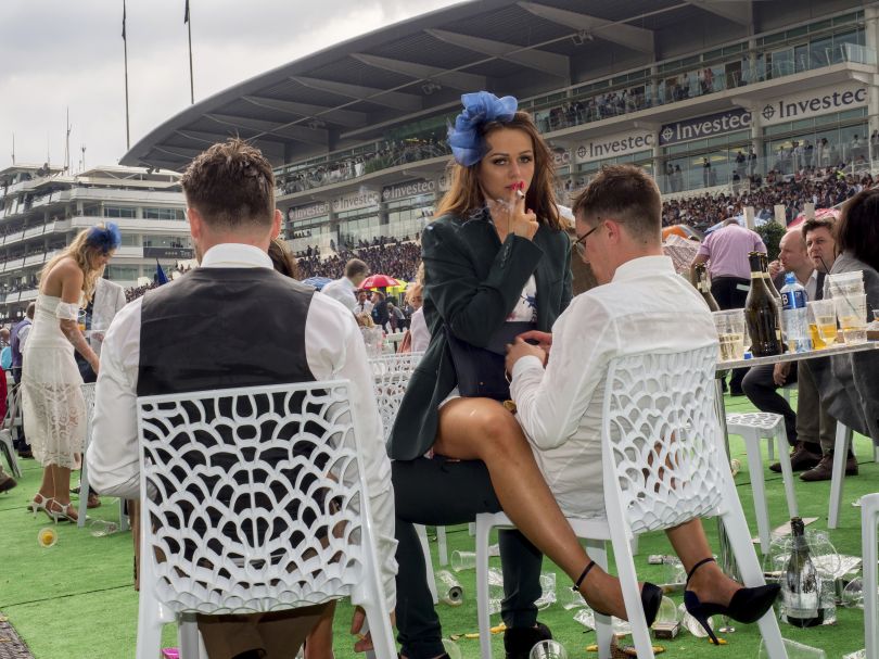 A young woman smokes a cigarette straddled across a man's lap on Ladies Day at Epsom. June 2017 © Peter Dench